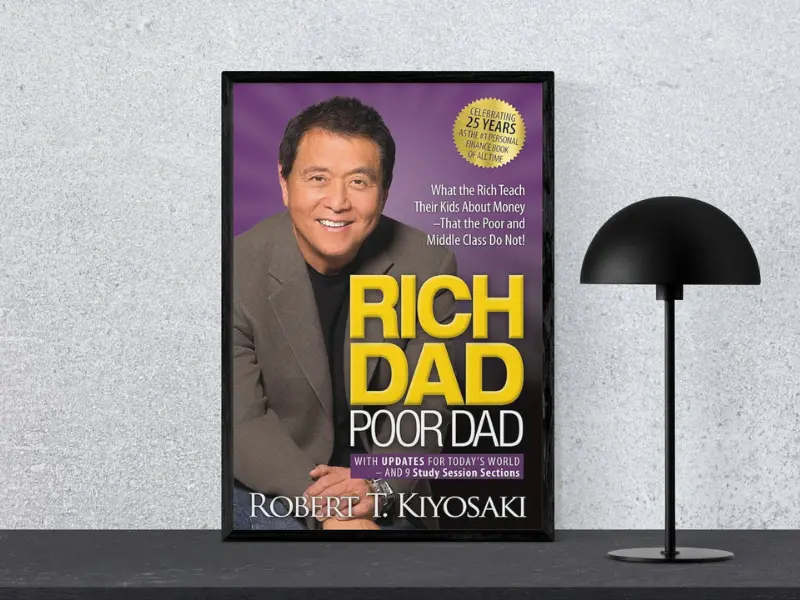 Book cover of "Rich Dad Poor Dad by Robert Kiyosaki, featuring the subtitle ‘Tiny Changes Remarkable Results’, displayed in a black frame next to a modern black lamp, a classic among the greatest business books