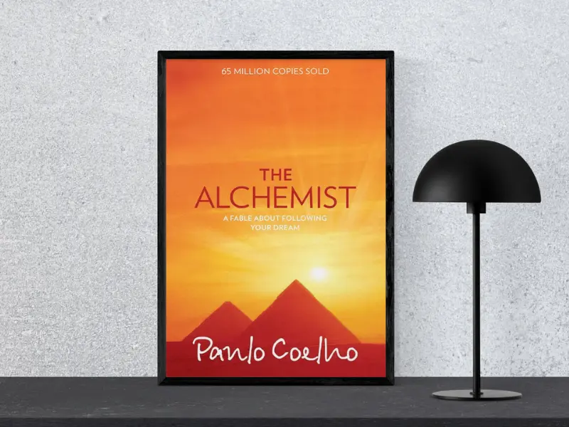 Book cover of "The Alchemist by Paulo Coelho, featuring the subtitle ‘Tiny Changes Remarkable Resutls’, displayed in a black frame next to a modern black lamp.