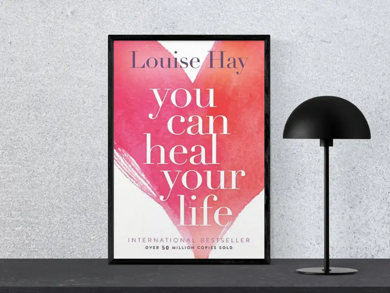 Book cover of "You Can Heal Your Life by Louise Hay, featuring the subtitle ‘Tiny Changes Remarkable Resutls’, displayed in a black frame next to a modern black lamp.