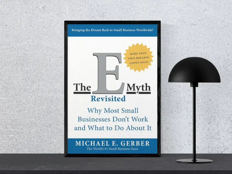 Book cover of "The E-Myth Revisited by Michael E. Gerber, featuring the subtitle ‘Tiny Changes Remarkable Results’, displayed in a black frame next to a modern black lamp, from the collection of greatest business books.