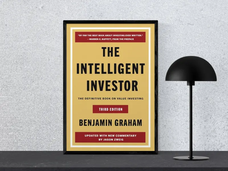 Book cover of "The Intelligent Investor by Benjamin Graham, featuring the subtitle ‘Tiny Changes Remarkable Results’, displayed in a black frame next to a modern black lamp, highlighting one of the greatest business books.