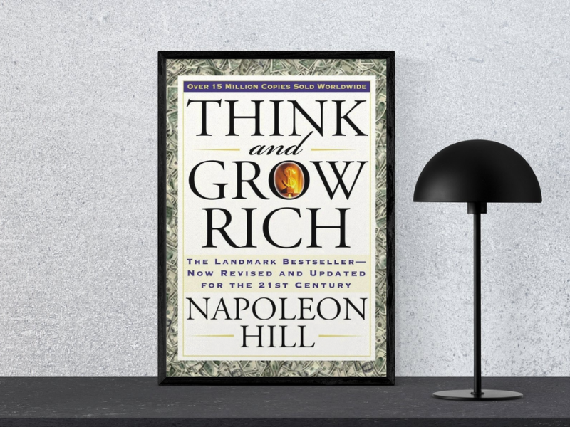 Book cover of "Think and Grow Rich by Napoleon Hill, featuring the subtitle ‘Tiny Changes Remarkable Resutls’, displayed in a black frame next to a modern black lamp.