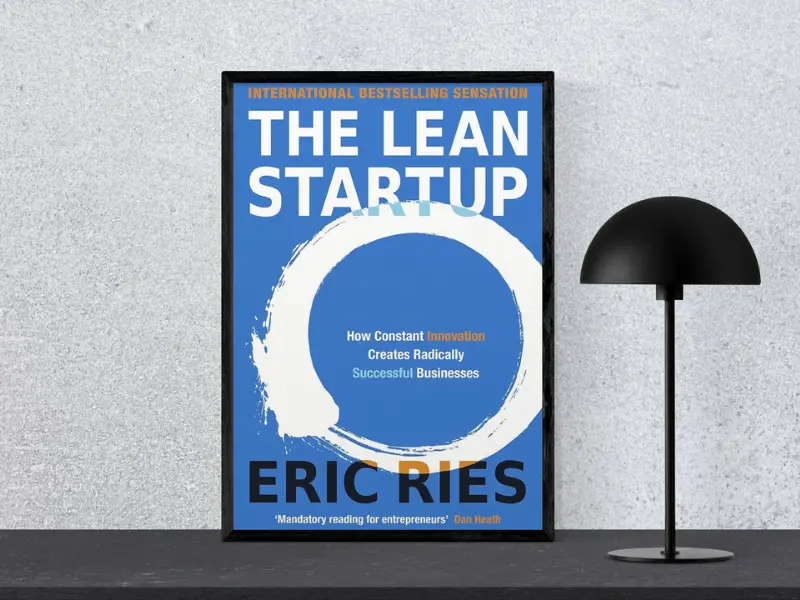 Book cover of "The Lean Startup by Eric Ries, featuring the subtitle ‘Tiny Changes Remarkable Results’, displayed in a black frame next to a modern black lamp, counted among the greatest business books.
