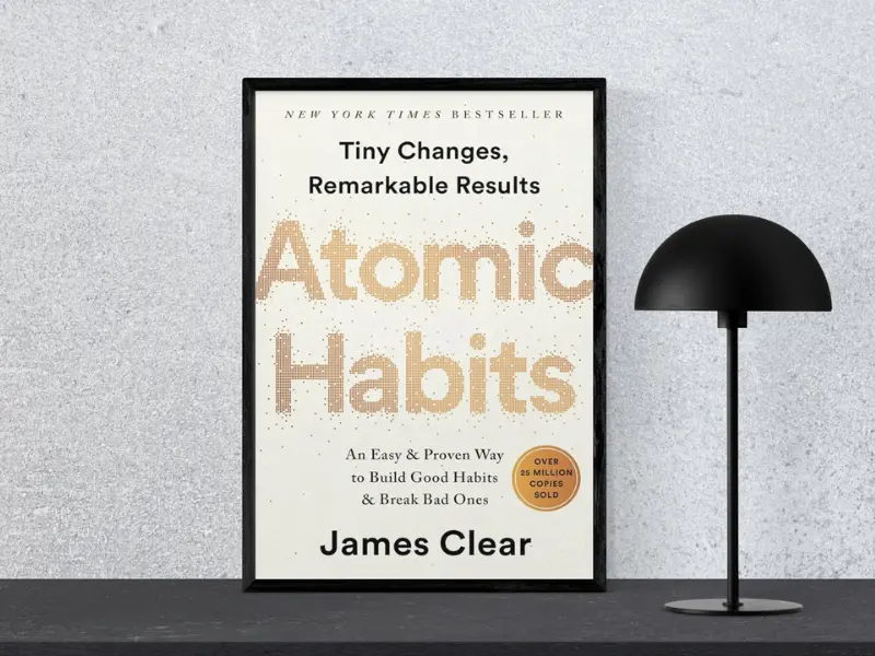 Atomic Habits Book Review Book cover of "Atomic Habits by James Clear, featuring the subtitle ‘Tiny Changes Remarkable Resutls’, displayed in a black frame next to a modern black lamp.