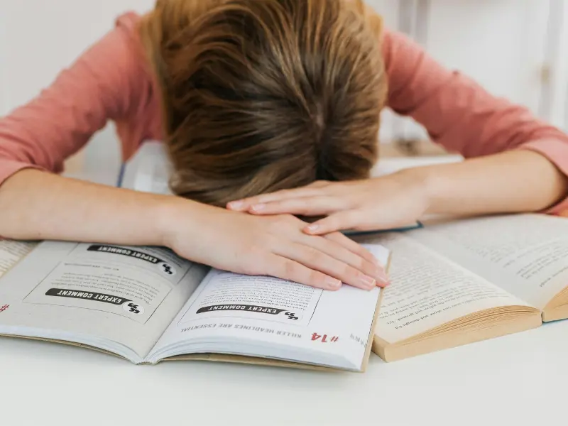 “Indian student wearing an orange t-shirt with head down on a study table, showing exhaustion and sadness. The image reflects the crisis of student mental health in India, emphasizing stress, anxiety, and pressure from studies.”